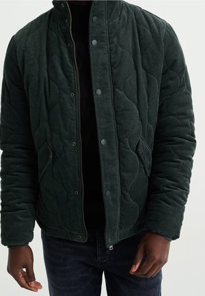 Mand iført mørkegrøn, quiltet kordfløjlsjakke med trykknapper og lynlås, kombineret med mørke jeans og sort skjorte under.