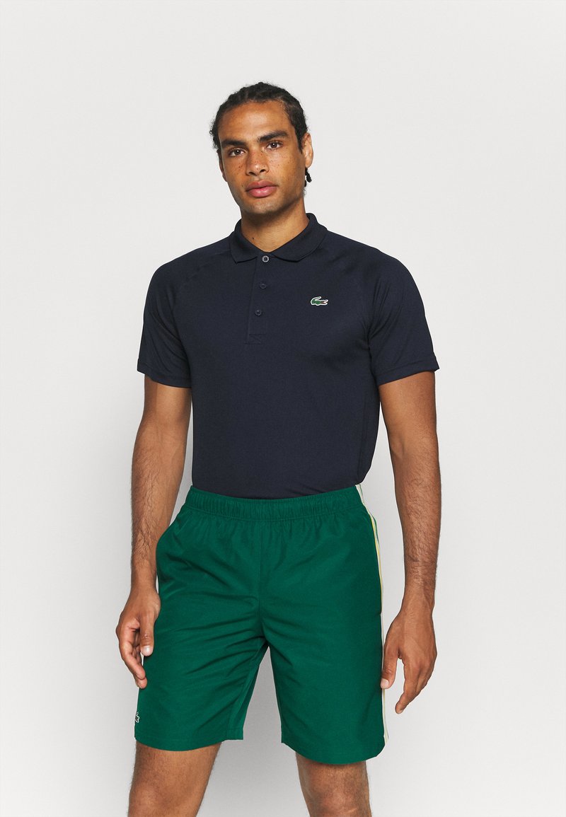 Camisa polo azul marino con cuello y tres botones, combinada con pantalones cortos verdes con franjas laterales. La tela parece suave y ligera.