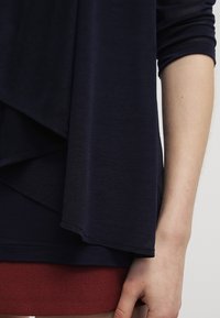 Navy Langarmshirt mit drapiertem Vorderteil, weichem Stoff, abgerundetem Saum und einem schichtartigen Look über einem roten Kleidungsstück.
