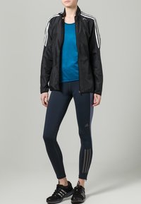 Black zip-up athletic jacket with white stripes, paired with a teal shirt and navy leggings featuring a grey stripe. Black athletic shoes complete the outfit.