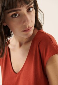 Top met V-hals in roestkleur met korte mouwen, gemaakt van gladde stof. De halslijn en mouwen hebben een strak, eenvoudig ontwerp zonder versieringen.
