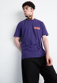Purple crew-neck t-shirt made of cotton, featuring a red "Carhartt" logo on the left chest. Model wears black pants, leaning against a wall.