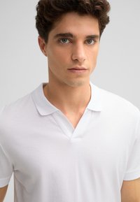 Weißes Baumwoll-Polohemd mit einem kurzen kragenlosen Ausschnitt und einem V-Ausschnitt-Design, das eine glatte Textur und eine taillierte Silhouette aufweist.