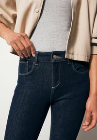 Los pantalones vaqueros de denim azul oscuro presentan una cintura alta, bolsillos clásicos y costuras claras en contraste. Se combinan con una blusa gris clara y una chaqueta beige.
