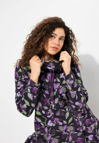 Sudadera con estampado floral en tonos púrpura y verde, con un cuello alto, detalles de cordones y tejido suave. La modelo ajusta el cuello.