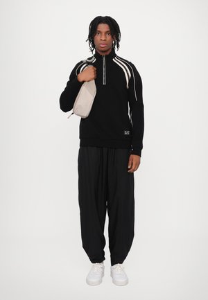 Black zip-up sweater with white stripes, paired with black wide-leg pants and white sneakers. The model holds a beige sling bag.