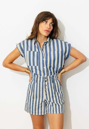 Femme portant une chemise à manches courtes rayée verticale bleue et blanche et un short taille haute assorti, debout les mains sur les hanches sur fond blanc.