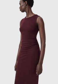 Burgundin värinen hihaton mekko, valmistettu pehmeästä kankaasta, jossa on vartalonmyötäinen siluetti ja sivulla laskostettu yksityiskohta.