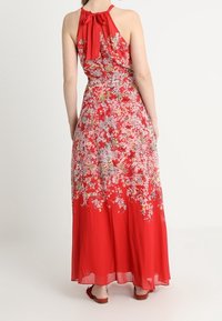 Robe maxi fleurie rouge à encolure halter avec un nœud, en tissu léger et transparent, silhouette évasée avec des motifs de fleurs roses et jaunes.