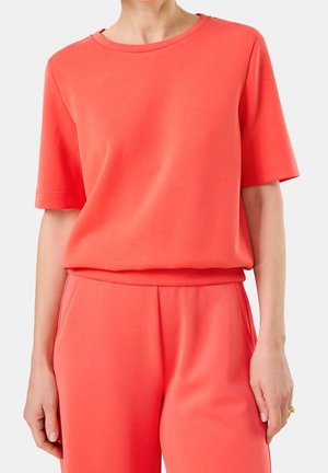 Een vrouw draagt een koraalkleurig shirt met korte mouwen en een bijpassende losse broek, met haar handen langs haar zijkanten tegen een effen achtergrond.