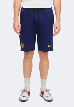 Muž oblečený v námornícky modrých športových krátkych nohaviciach so žltými logami, sivých ponožkách a bielych a červených teniskách Nike stojí pred jednoduchým pozadím.