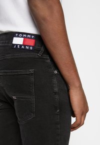 Persona vistiendo pantalones negros de Tommy Jeans con parche de la marca visible en la cintura y el bolsillo trasero, camisa blanca metida dentro.