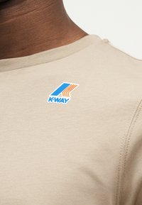 Λαιμός μπεζ πουκαμίσου με τυπωμένο μπλε και πορτοκαλί λογότυπο K-Way κοντά στον γιακά στο πάνω μέρος του στήθους ενός ατόμου.