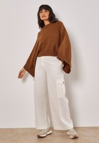 Brauner Strickpullover mit weiten Ärmeln, kombiniert mit einer locker sitzenden weißen Cargo-Hose mit Seitentaschen und beigen Sneakers.
