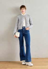 Knit gray cardigan with gold buttons, white shirt underneath, paired with blue flared jeans and chunky white sneakers. Minimalist white handbag included.