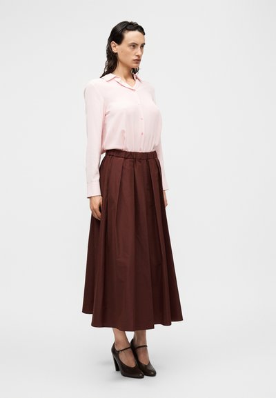 Ανοιχτό ροζ μπλούζα με μακριά μανίκια, κλείσιμο με κουμπιά και γιακά, συνδυασμένη με φούστα ψηλόμεση, ρέουσα, σκούρου καφέ χρώματος. Καφέ παπούτσια με τακούνι.