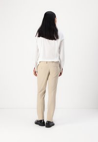Pantaloni beige su misura realizzati in tessuto liscio, con vestibilità slim e due tasche posteriori. Abbinati a una camicia bianca a maniche lunghe e scarpe nere.