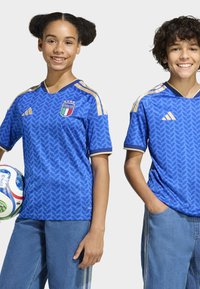 Blå fotbollströjor med ett lövmönster, gulddetaljer och Italiens emblem. Matchas med ljusblå jeans, en håller i en fotboll.