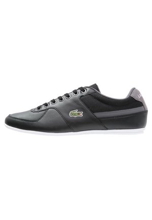 Czarny niski sneaker z wiązaniem na sznurowadła, biała podeszwa, szara zapiętka i zielone logo krokodyla po boku.