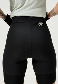 Een persoon is van achteren te zien, gekleed in zwarte fietsbroekjes met padding en een grijs shirt, met een logo op de taille.