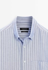 Jasnoniebieska koszula w paski z kołnierzykiem zapinanym na guziki, białymi pionowymi paskami oraz kieszonką na lewej piersi. Wykonana z materiału bawełnianego.