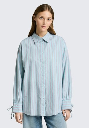 Młoda kobieta o długich włosach nosi oversize'ową, jasnoniebieską koszulę na guziki w pionowe paski z zawiązanymi mankietami, do tego niebieskie dżinsy.