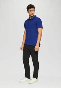 Blaues Polohemd mit Kragen und kurzen Ärmeln, kombiniert mit schwarzen Hosen und weißen Sneakers. Der Stoff wirkt glatt und enganliegend.