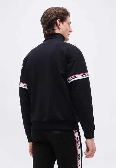 Άνδρας που φοράει μαύρο φόρμα Moschino με λευκές και κόκκινες ραμμένες λωρίδες στα μανίκια και στις πλευρές των παντελονιών, με την πλάτη προς τα εμπρός σε απλό φόντο.