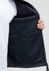 Interior de chaqueta azul marino con cremallera, fabricado con tejido texturizado; presenta un bolsillo y un ajuste en el dobladillo con un cordón. Detalle de costura visible.