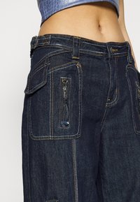Denim cargo pants in dark blue with multiple pockets. Contrast yellow stitching, zippered side pocket, and a classic waistband design.