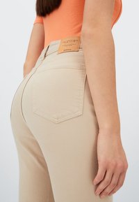 Kvinna i beige mom slim jeans och orange kortärmad topp, sedd bakifrån från midjan till mitten av låren med en avslappnad hand vid sidan.