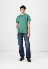 Grønn bomull T-skjorte med korte ermer, rund hals, og liten logo på brystet, kombinert med blå denimjeans og sorte sko.