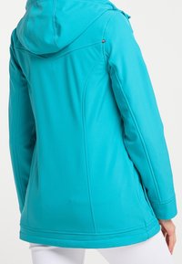 Turkos huva mjukskaljacka med långa ärmar, bärs av en person som visas bakifrån med vita byxor synliga.