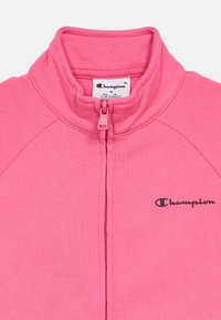 Ροζ fleece μπουφάν με ψηλό λαιμό, μπροστινό φερμουάρ και μαύρο λογότυπο "Champion". Διαθέτει ραφές ράγ κλαν και μαλακή υφή. Μέγεθος S.