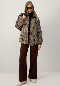 Barvita jakna s paisley vzorcem in ovratnikom, kombinirana s širokimi rjavimi hlačami in belimi supergami, nošena s črnimi sončnimi očali.