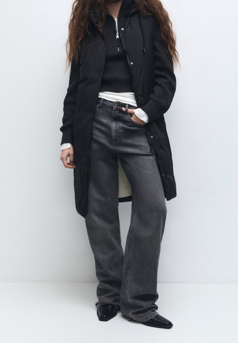 Personne portant une longue veste noire sans manches, un pull noir sur une chemise blanche, un jean large gris foncé et des chaussures noires pointues.