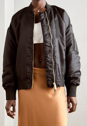 Person wearing unzipped black bomber jacket over white crop top and tan skirt, accessorized with gold necklace and rings on both hands.