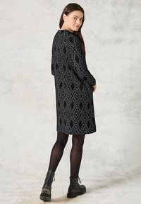 Svart klänning med diamantmönster, långa ärmar och rund halsringning; matchad med svarta ankelboots och svarta strumpbyxor. Slät tygstruktur.