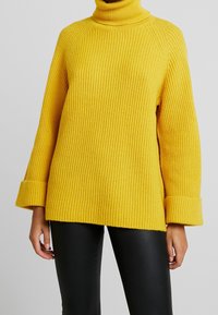 Žlutý žebrovaný rolák s dlouhými rukávy a širokými manžetami. Tkanina má texturovaný povrch, což poskytuje útulný vzhled.