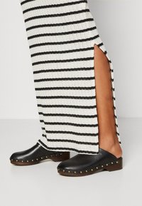 Sabots en cuir noir avec des clous dorés le long de la semelle, portés avec une robe en tricot rayée blanche et noire avec des fentes sur les côtés.