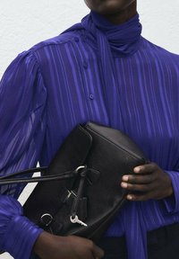 Borsa nera in pelle con finitura testurizzata, ferramenta argentata e tracolla, indossata sopra una camicia viola a righe.