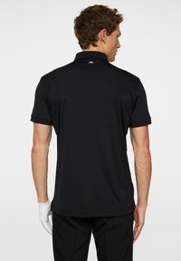 Svart polo skjorta sedd bakifrån, med klassisk krage, korta ärmar och en liten logotyp vid nacken. Slät tygtextur.