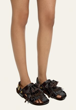 Stopy w ciemnobrązowych skórzanych sandałach z odkrytymi palcami, z warstwowymi frędzlami i chwostami, zapinane na paski z złotą klamrą.