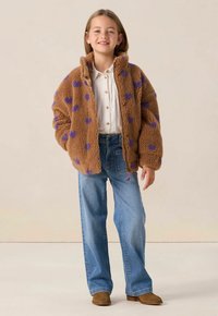 Veste brune et pelucheuse avec des motifs de cœurs violets, portée avec un jean large bleu et des bottines brunes. Chemise blanche à boutons en dessous.