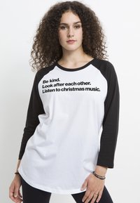 Vit långärmad skjorta med svarta raglanärmar, med fet svart text som säger "Var snäll. Ta hand om varandra. Lyssna på julmusik."