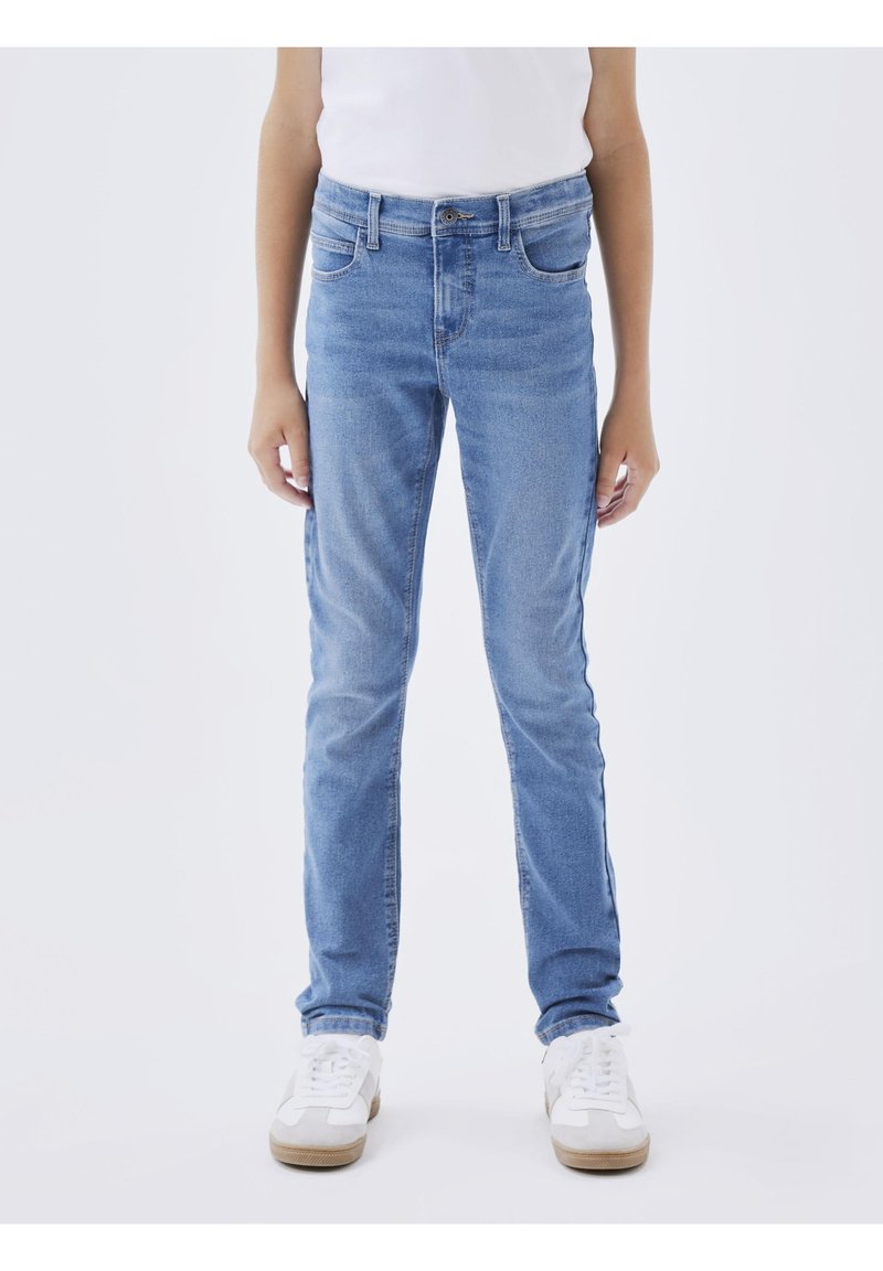 Personne portant un jean bleu clair à jambe droite, des baskets blanches et un top blanc sans manches, debout avec les bras détendus le long du corps.
