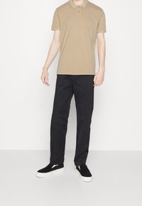 Polo beige avec une patte de boutonnage à deux boutons, associé à un pantalon noir et des baskets noires à enfiler avec une semelle blanche. Design minimaliste.