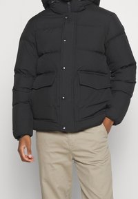 Czarna kurtka puchowa z kapturem, zatrzaskami i dwoma przednimi kieszeniami. Materiał ma gładką fakturę i umiarkowaną izolację.