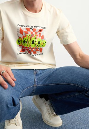 Person trägt cremefarbenes Adidas T-Shirt mit orange-grünem Motiv, blaue Jeans, weiße Sneaker, sitzt im Schneidersitz auf blauem Boden.