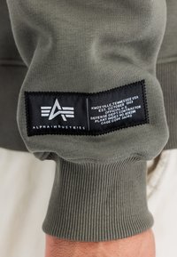 Sweatshirt verde oliva con polsini e orlo a costine. Presenta un'etichetta nera con testo e logo bianchi, indicanti i dettagli del marchio e della produzione.